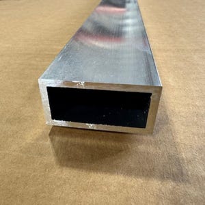 May include: A rectangular aluminum tube with a hollow black interior. The metal has a brushed finish and is resting on a brown surface. The tube's dimensions are not visible.