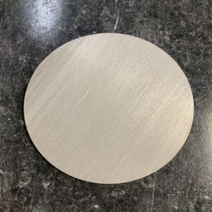 May include: A round, light gray object with a textured surface. The object is circular and appears to be made of a solid material. The surface has a subtle, grainy pattern. The object is set against a dark, mottled background.