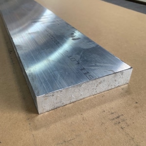 May include: A rectangular, brushed aluminum metal bar. The bar has a matte finish and is resting on a brown surface. The text "LOT 101 115" is printed on the bar. The bar is likely used for construction or manufacturing.