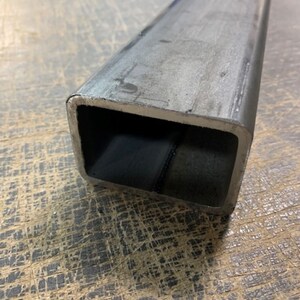 May include: Close-up of a square-shaped metal tube. The tube is made of steel and has a silver finish. The tube is lying on a textured surface.