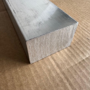 May include: A rectangular, silver-colored metal bar with a brushed finish. The bar is shown in a close-up shot, highlighting its industrial texture and shape. The bar is likely used for construction or manufacturing purposes.