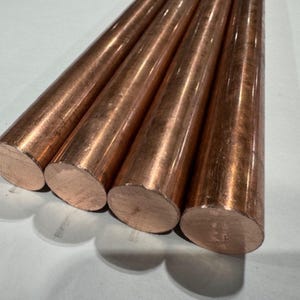May include: Four copper rods with a polished, reflective surface. The rods are cylindrical with flat, cut ends, displaying a warm, reddish-brown color. The rods are arranged diagonally, casting shadows on a light-colored surface.