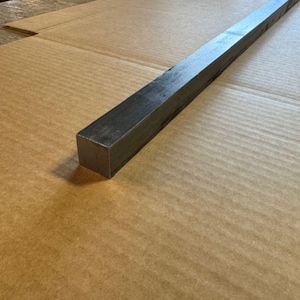 May include: A long, rectangular piece of metal with a square cross-section. The metal is a dark gray color and has a smooth, polished finish.