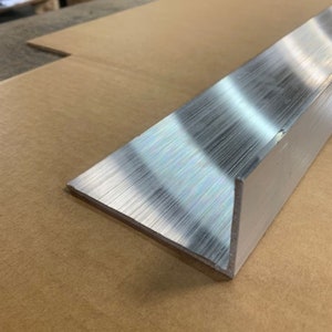 May include: A silver aluminum corner guard with a brushed finish. The corner guard is on a brown cardboard surface.