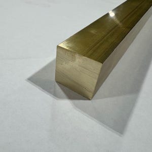 May include: A solid brass square bar with a polished finish. The bar is rectangular in shape and casts a shadow on the white surface. The brass has a golden color.