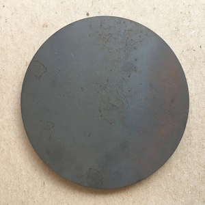 Puede incluir: Un disco redondo de metal gris oscuro con una superficie ligeramente texturizada. El disco tiene un aspecto desgastado y envejecido con algunas manchas de &oacute;xido.