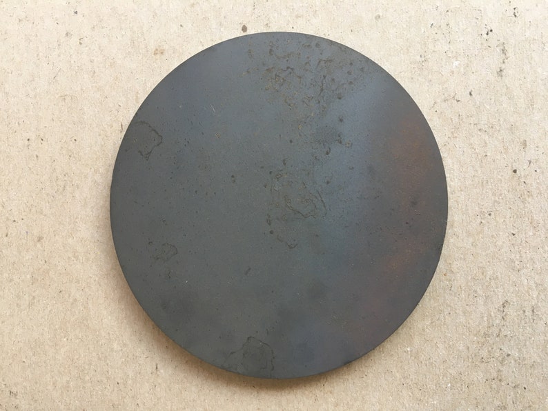 Puede incluir: Un disco redondo de metal gris oscuro con una superficie ligeramente texturizada. El disco tiene un aspecto desgastado y envejecido con algunas manchas de &oacute;xido.