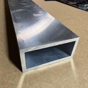 May include: A long, rectangular aluminum extrusion with a hollow, rectangular cross-section. The extrusion has a brushed finish and is lying on a brown surface.