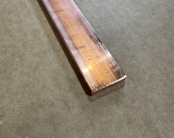 May include: A rectangular copper bar with a polished surface. The bar is a reddish-brown color and appears to be made of solid metal. The bar is lying on a neutral-colored surface.