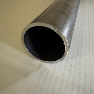 May include: A close-up view of a cylindrical metal tube. The tube is silver-colored with a dark interior. The end of the tube shows a rough, cut edge. The tube is lying on a light-colored surface.
