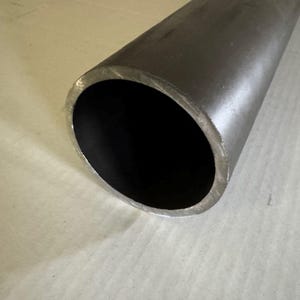 May include: A close-up of a cylindrical metal tube with a dark interior. The tube is a dark gray color and appears to be made of steel. The end of the tube is visible, showing the hollow interior.