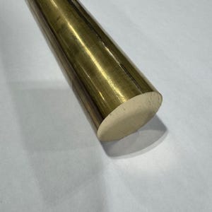 May include: A solid brass rod with a rounded end. The rod has a polished, golden finish and is cylindrical in shape. The end is a lighter, unpolished brass color. The rod is lying diagonally across a white surface.