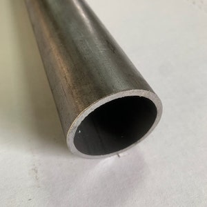 May include: A cylindrical metal tube with a dark gray, slightly textured surface. The tube has a hollow interior and a smooth, rounded edge. The metal appears to be unfinished or raw, with subtle variations in color and texture.