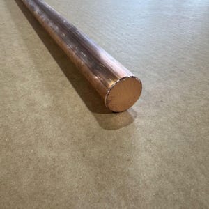 May include: A copper rod with a cylindrical shape, lying diagonally across a neutral-toned surface. The rod has a polished, metallic finish with a visible end face. The item is likely used for construction or crafting.