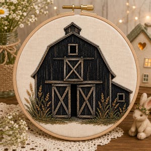 May include: A circular wooden embroidery hoop containing a fabric panel with a hand-stitched design of a dark charcoal barn. The barn features a central sliding door, a small upper window, and a cupola. Stitched wheat stalks flank the structure.