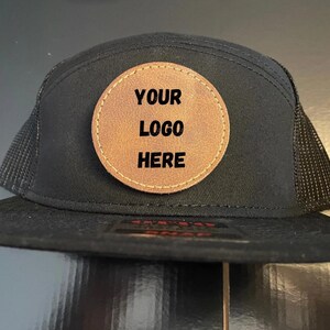 May include: Black trucker hat with a brown leather patch that says "YOUR LOGO HERE" in black text.
