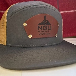 Op de afbeelding: Een grijze en beige baseballpet met een bruin leren patch waarop "NGU NEVER GIVE UP EST. 2020" staat met een berg en een silhouet van een persoon.