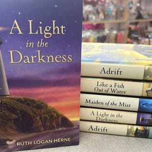 May include: A stack of books with titles including "A Light in the Darkness" and "Adrift." The cover of "A Light in the Darkness" features a lighthouse on a hill at sunset. Other books have scenic covers.