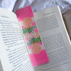 May include: A rectangular bookmark with a bright pink background featuring two peach-coloured checkered vases filled with pink and green floral arrangements. The top of the bookmark displays the text "I am here" with small arrows.