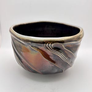 Pode incluir: Uma tigela de cerâmica com um interior profundo e escuro e um esmalte exterior brilhante de vários tons. A superfície apresenta padrões de redemoinho em cobre metálico, bronze e cinzento-carvão, com cristas texturizadas no corpo.