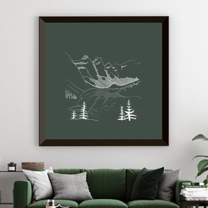 May include: A framed art print with a white line drawing of a mountain scene on a dark green backdrop. The artwork depicts mountains, trees, and a lake. The frame is dark brown. The print is displayed above a green sofa.