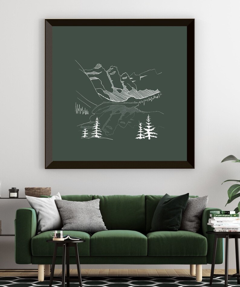 May include: A framed art print with a white line drawing of a mountain scene on a dark green backdrop. The artwork depicts mountains, trees, and a lake. The frame is dark brown. The print is displayed above a green sofa.