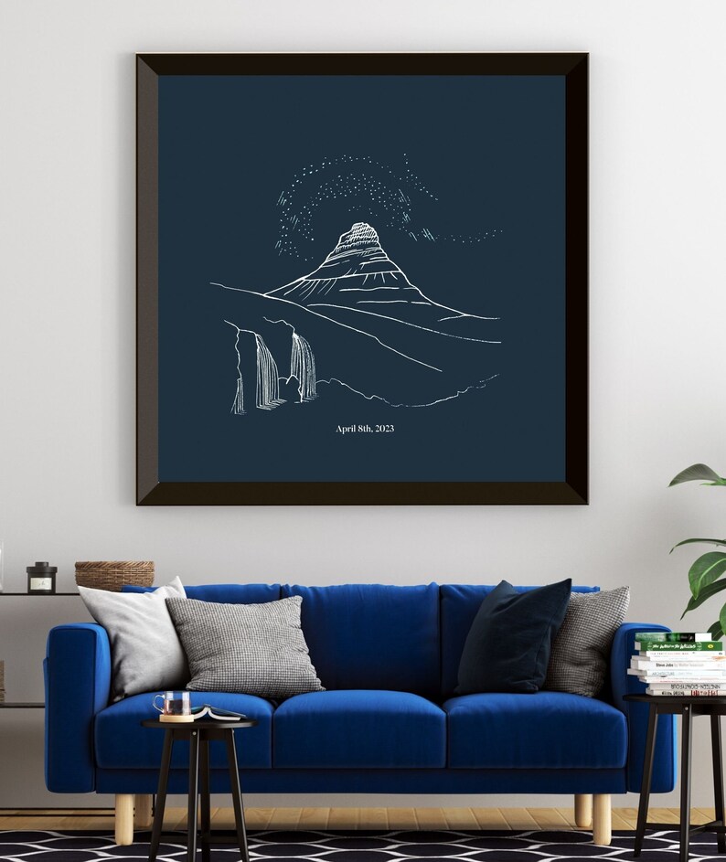 May include: Framed art print featuring a white line drawing of a mountain landscape with a waterfall and starry sky on a navy blue background. The date "April 8th, 2023" is printed at the bottom. The artwork is displayed above a blue sofa.