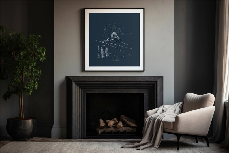 May include: A framed art print featuring a minimalist white line drawing of a mountain landscape on a dark blue background. The artwork is displayed above a black fireplace, with a beige armchair and a potted plant in the room.