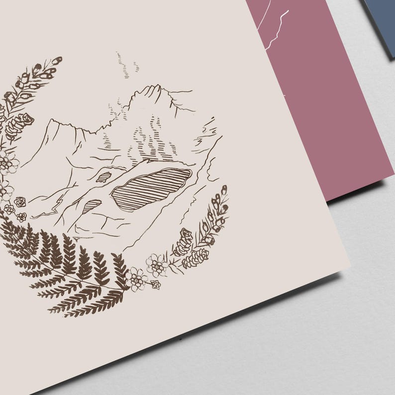 May include: A print featuring a mountain landscape illustration in brown ink on a light beige background. The mountain scene is surrounded by a wreath of ferns and flowers. Other prints in shades of pink and blue are visible in the background.