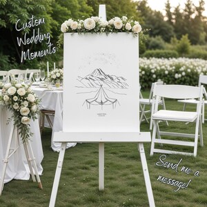 May include: A white wedding sign on an easel, decorated with flowers. The sign displays a mountain and tent illustration, with the text "Custom Wedding Moments" and "Send me a message!". The setting is outdoors, with white chairs and a table visible.