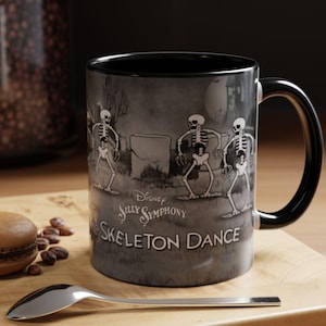 May include: Black ceramic mug with a grayscale illustration of dancing skeletons from Disney's Silly Symphony "The Skeleton Dance." The mug has a black interior and handle. A spoon and a pastry are on the wooden surface.