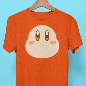 Könnte beinhalten: Orangefarbenes T-Shirt mit einem Comic-Charaktergesicht auf der Vorderseite. Der Charakter hat große Augen und einen rosa Rouge.
