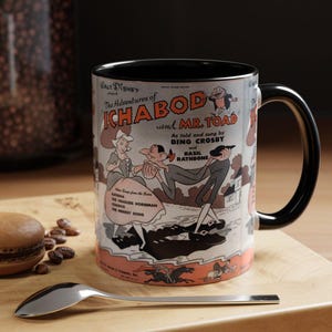 May include: A ceramic mug with a vintage design from Walt Disney's "The Adventures of Ichabod and Mr. Toad." The mug has a black handle and interior, with a colourful illustration and text from the animated film. A spoon and a macaron are on the wooden surface.