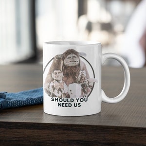 May include: White ceramic mug with a graphic of the characters from the movie Labyrinth. The graphic shows a furry creature with a human-like face, a small human-like creature, and a small white creature. The text "Should You Need Us" is printed below the graphic.