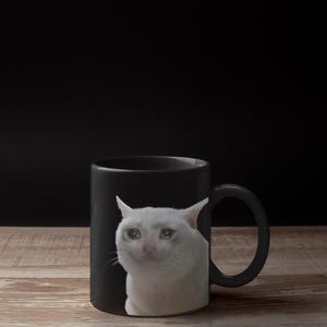 Puede incluir: Taza de cerámica negra con un gato blanco con una expresión triste y lágrimas. La imagen del gato está impresa en la parte delantera de la taza. La taza tiene un asa negra y está sobre una superficie de madera.