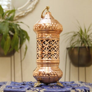May include: A copper-colored lantern with an ornate design. The lantern has a handle at the top and sits on three small feet. The middle section features a lattice pattern. The lantern is placed on a blue patterned cloth.