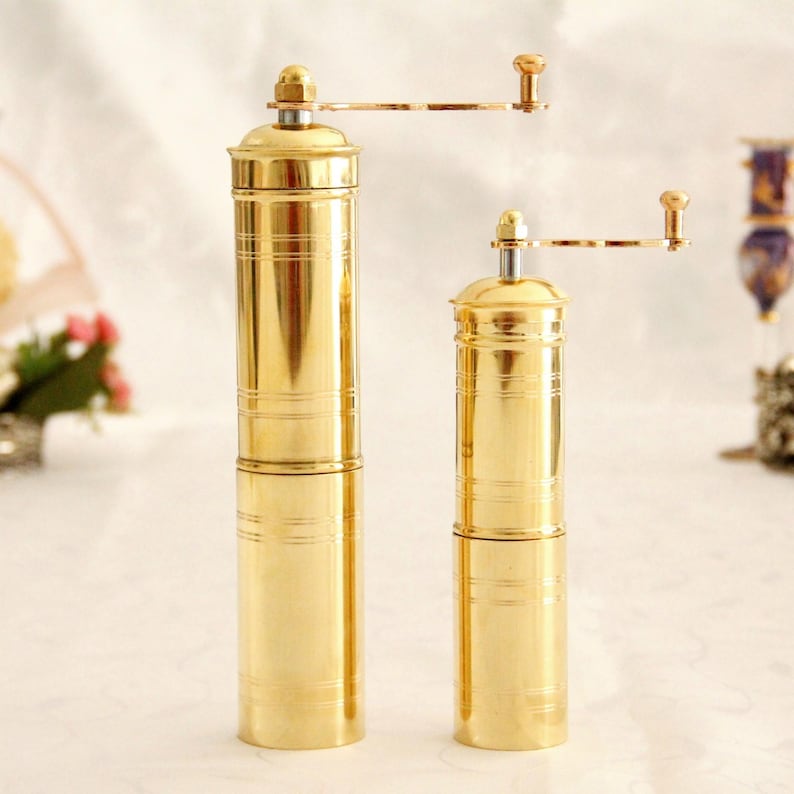 May include: Two gold manual coffee grinders with a crank handle. The grinders are made of brass and have a cylindrical shape. The larger grinder is on the left and the smaller grinder is on the right.