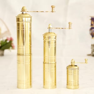 May include: Three gold metal pepper grinders of different sizes. The grinders have a crank handle and are made of polished brass.