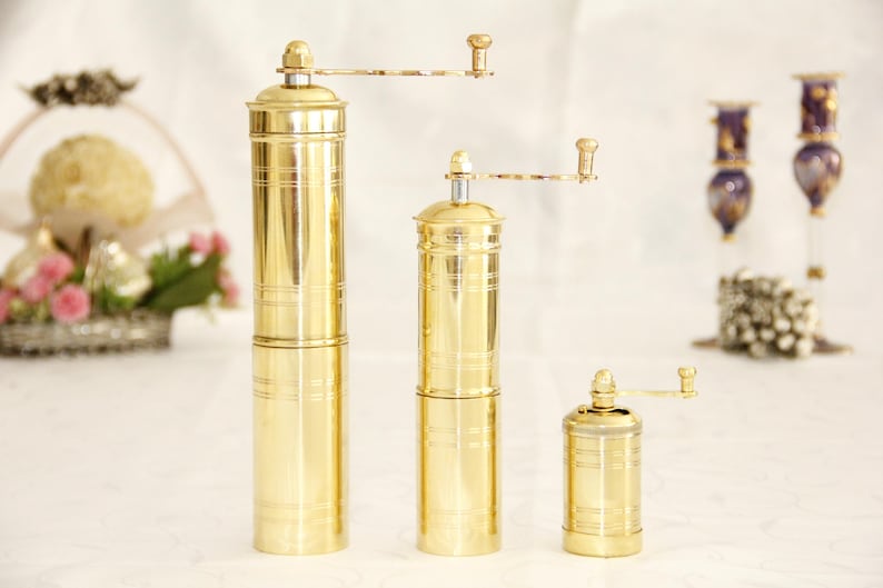 May include: Three gold metal pepper grinders of different sizes. The grinders have a crank handle and are made of polished brass.