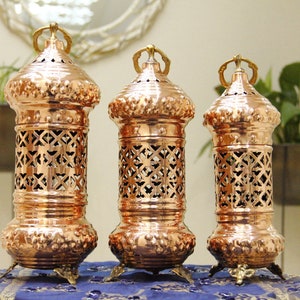 May include: Three copper lanterns with intricate cut-out designs. Each lantern has a cylindrical body, a domed top with a handle, and ornate feet. The lanterns are arranged on a blue patterned surface, with greenery in the background.