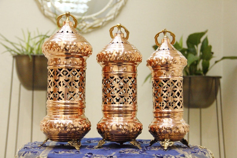 May include: Three copper lanterns with intricate cut-out designs. Each lantern has a cylindrical body, a domed top with a handle, and ornate feet. The lanterns are arranged on a blue patterned surface, with greenery in the background.