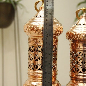 May include: Two copper-colored decorative lanterns, approximately 12 inches tall, with intricate cut-out patterns and ornate detailing. Each lantern has a handle and sits on small feet. A ruler is placed next to the lantern.