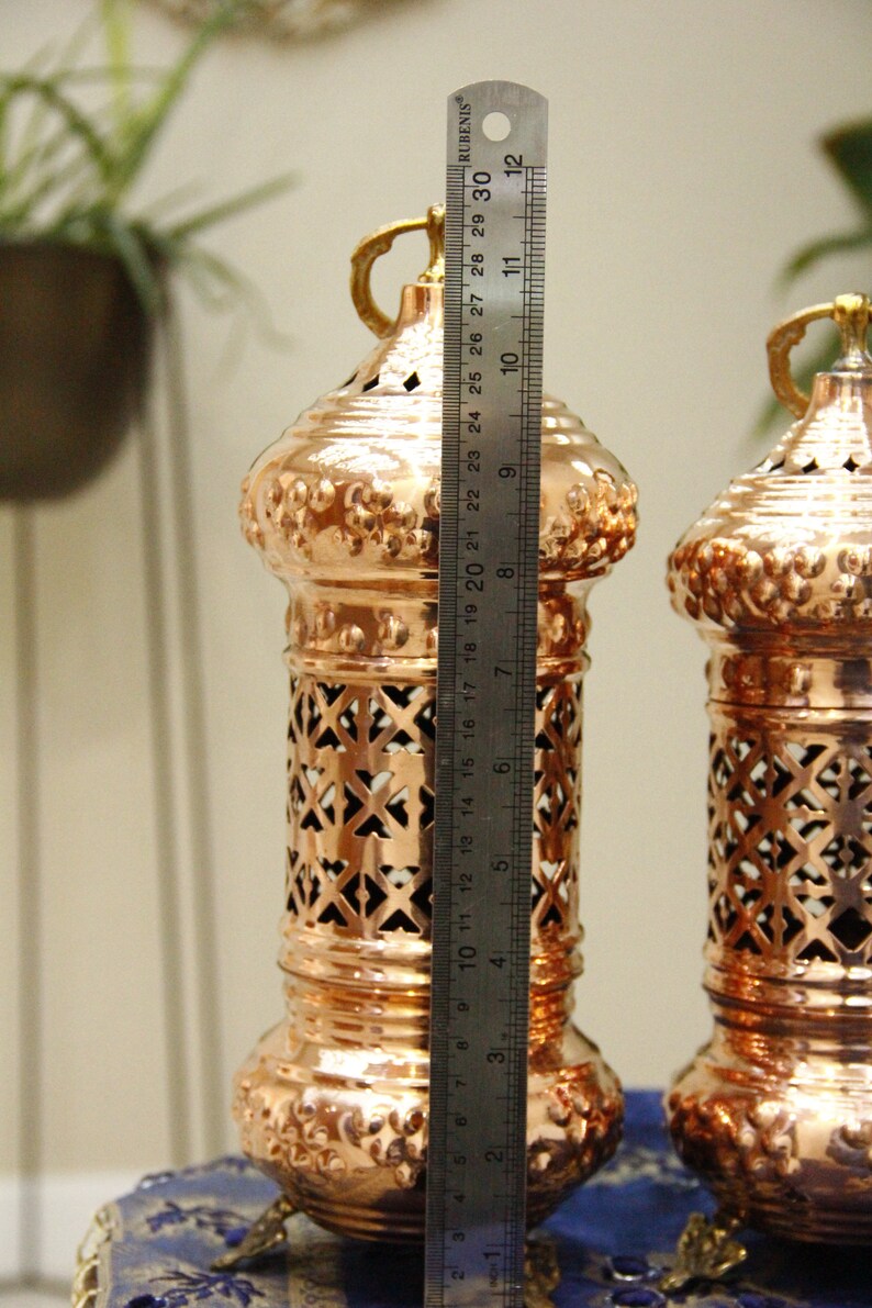 May include: Two copper-colored decorative lanterns, approximately 12 inches tall, with intricate cut-out patterns and ornate detailing. Each lantern has a handle and sits on small feet. A ruler is placed next to the lantern.