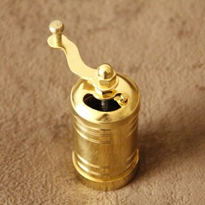 May include: A gold metal pepper grinder with a crank handle. The grinder has a cylindrical shape and a smooth, polished finish.