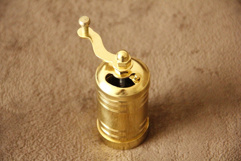 May include: A gold metal pepper grinder with a crank handle. The grinder has a cylindrical shape and a smooth, polished finish.