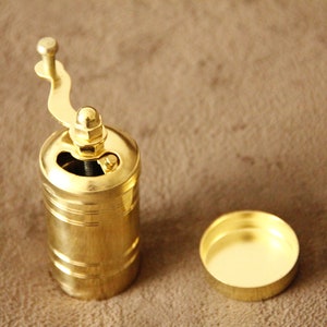 May include: A gold metal coffee grinder with a crank handle. The grinder is open and the top is off. The grinder is sitting on a brown surface.