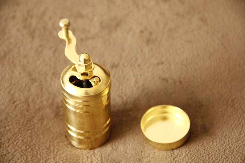 May include: A gold metal coffee grinder with a crank handle. The grinder is open and the top is off. The grinder is sitting on a brown surface.