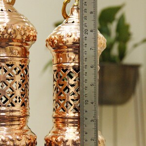 May include: Two copper-colored lanterns with intricate cut-out designs and ornate bases. Each lantern is approximately 10 inches tall, with a decorative handle at the top. The lanterns are displayed on a blue cloth.