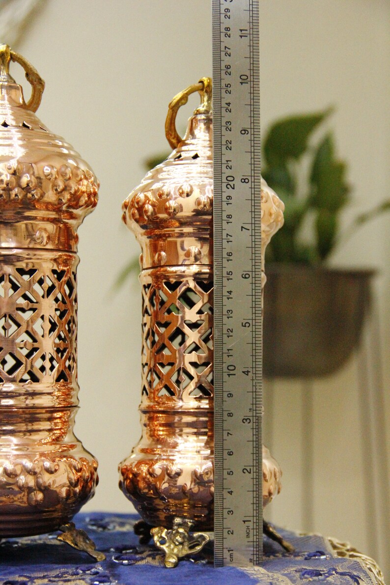 May include: Two copper-colored lanterns with intricate cut-out designs and ornate bases. Each lantern is approximately 10 inches tall, with a decorative handle at the top. The lanterns are displayed on a blue cloth.