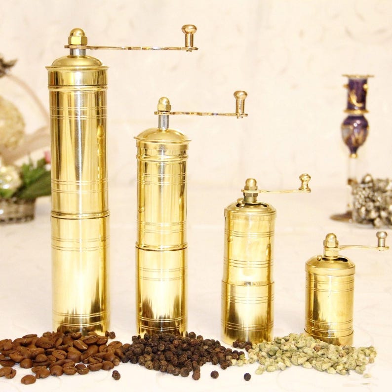 May include: Four gold-tone manual coffee grinders of varying sizes. The grinders are made of brass and have a traditional design. The grinders are arranged in a row from largest to smallest. Coffee beans, peppercorns, and green coffee beans are scattered in front of the grinders.